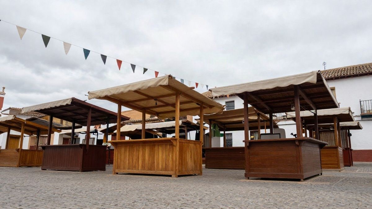 Imagen ilustrativa: puestos de madera con toldos en una plaza empedrada de un pueblo, con banderines y cielo nublado