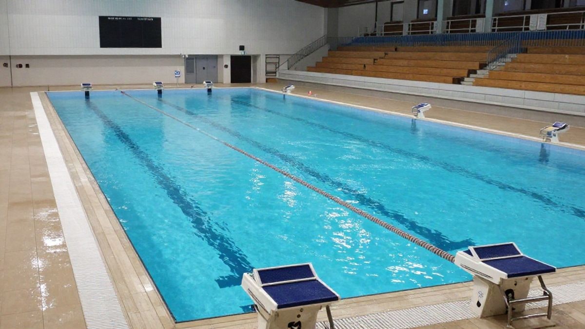 Illustrative image: empty indoor pool with lane ropes and starting blocks seen from the stands