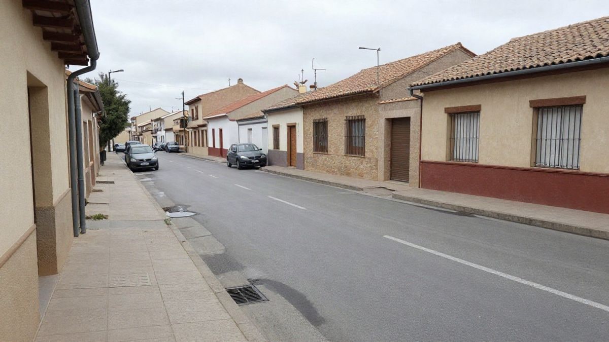 Imagen ilustrativa: calle residencial de un pueblo español con un sumidero y charcos bajo un cielo nublado