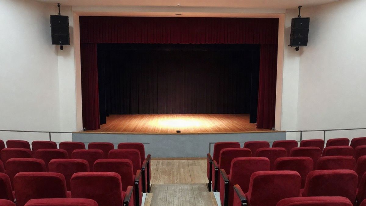 Imagen ilustrativa: interior de un teatro vacío con butacas rojas y un escenario con el telón cerrado, iluminado por un foco.
