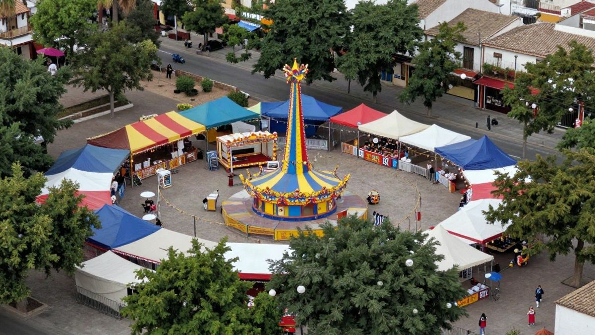 Imagen ilustrativa: vista cenital de un recinto ferial con atracciones y casetas en un parque urbano