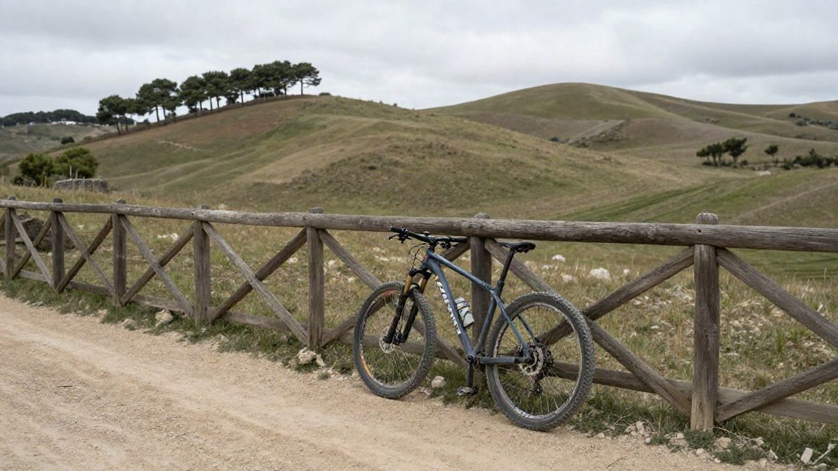 Illustrative image: mountain bike resting on a wooden fence next to a dirt path in a rural landscape with hills and pines in the background.
