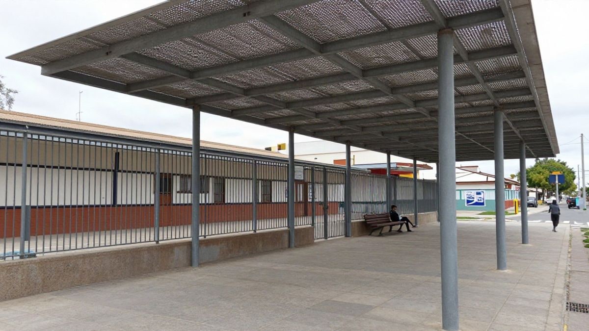 Imagen ilustrativa: pérgola metálica con cubierta microperforada sobre una acera frente a un colegio, con bancos y valla.