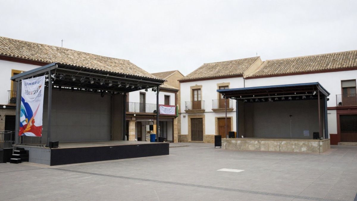 Imagen ilustrativa: plaza de un municipio español con escenario vacío y banderines, sin personas ni texto