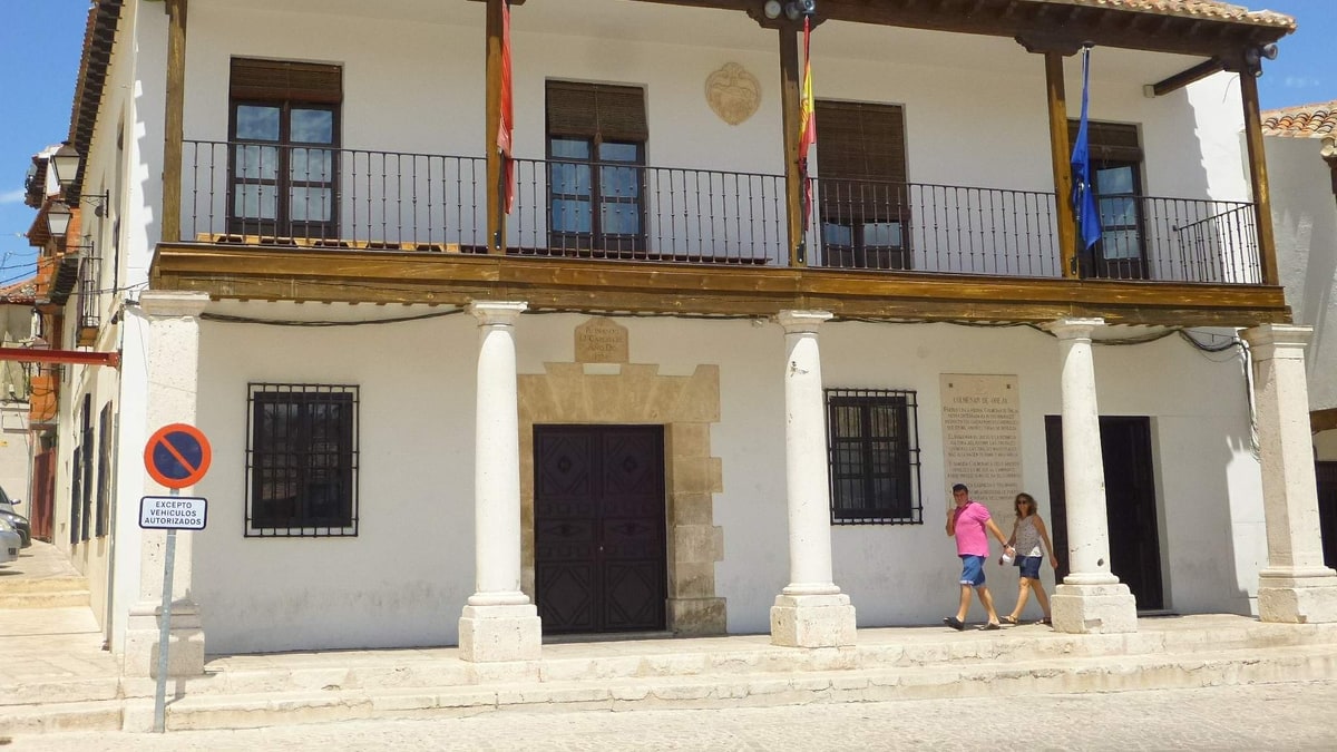 Fachada blanca del Ayuntamiento de Colmenar de Oreja con soportales, balcón de madera y dos personas paseando