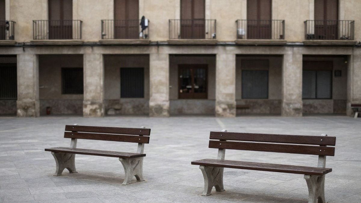Imagen ilustrativa: plaza municipal con bancos vacíos y edificio bajo al fondo en un día nublado