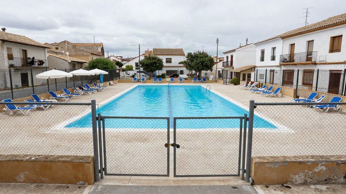 Imagen ilustrativa: una piscina municipal vacía con tumbonas y sombrillas, con la valla metálica cerrada con candado.