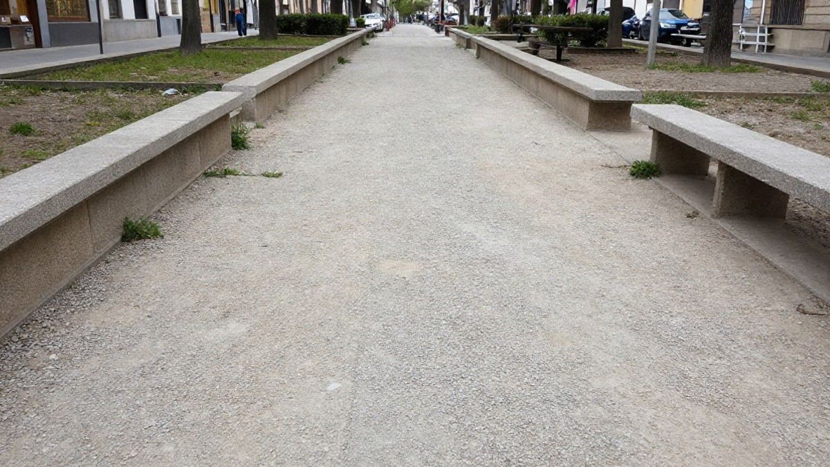 Imagen ilustrativa: sendero de grava en un parque urbano con muro bajo, bancos sencillos y hierba entre las juntas, bajo luz de día nublado