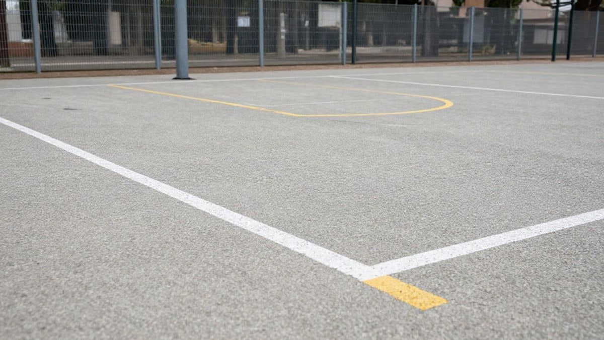 Imagen ilustrativa: primer plano del pavimento y las líneas de una pista multideportiva recién pintada, con portería y canasta al fondo