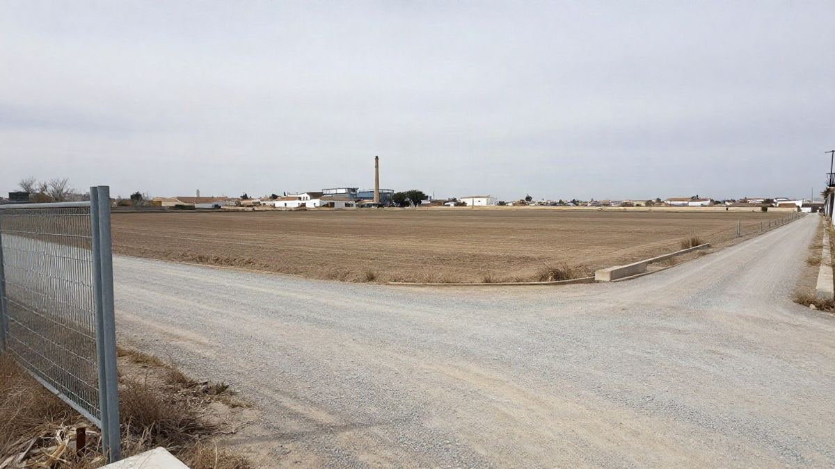 Illustrative image: flat and dry land on the outskirts of a municipality, with a generic industrial facility and a chimney in the background under a cloudy sky